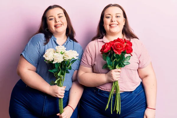 Young plus size twins holding flowers looking positive and happy standing and smiling with a confident smile showing teeth