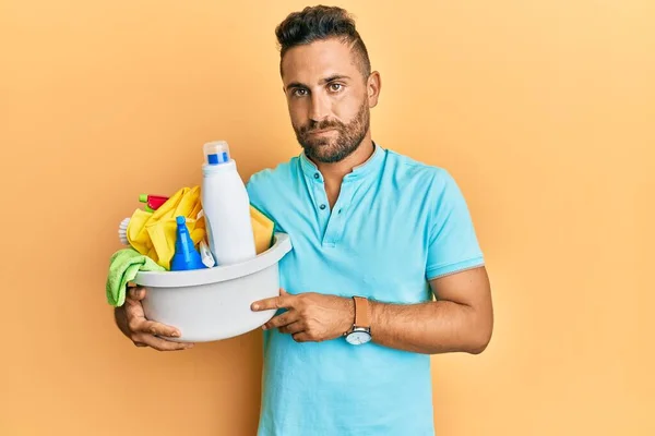 Handsome man with beard holding cleaning products thinking attitude and sober expression looking self confident