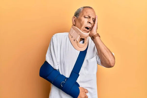 Handsome mature senior man wearing cervical collar and arm on sling yawning tired covering half face, eye and mouth with hand. face hurts in pain.