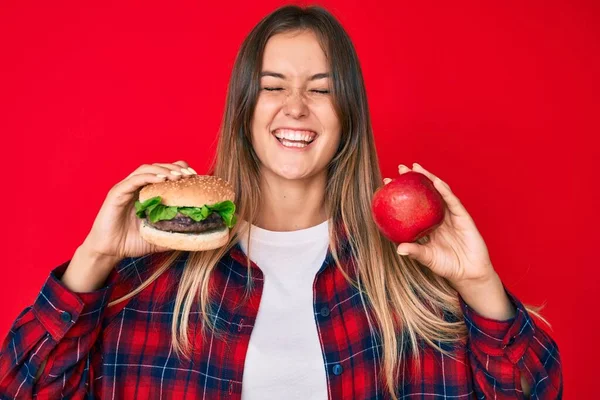 美丽的高加索女人比较汉堡包和健康的红苹果 笑得很开心 笑得很大声 因为有趣的疯狂笑话 — 图库照片