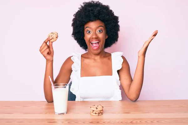 Young african american woman drinking glass of milk holding cookie celebrating victory with happy smile and winner expression with raised hands