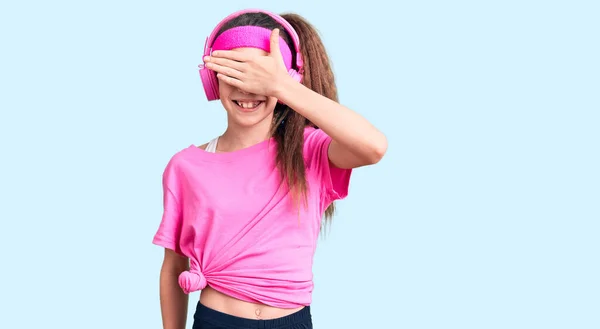 かわいいヒスパニック系の子供の女の子は ジムの服を着て 驚きのために目をカバー顔に手で笑ってヘッドフォンを使用しています 視覚障害の概念 — ストック写真