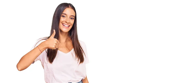 若いヒスパニック系の女性は手で幸せな親指アップジェスチャーを行うカジュアルな白いTシャツを着て 成功を示すカメラを見ると — ストック写真