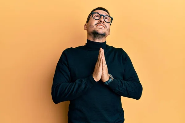 Handsome man with tattoos wearing turtleneck sweater and glasses begging and praying with hands together with hope expression on face very emotional and worried. begging.