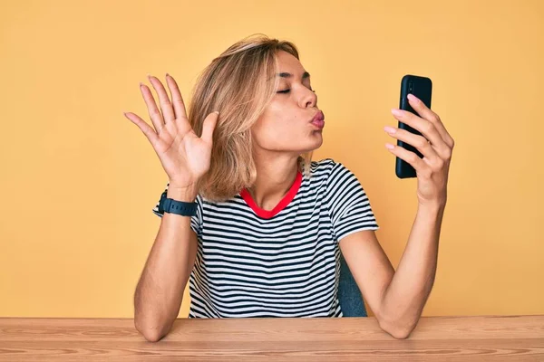 Beautiful caucasian woman doing video call waving to smartphone looking at the camera blowing a kiss being lovely and sexy. love expression.