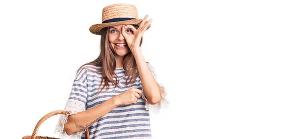 美しいCaucasian女性身に着けている夏の帽子と保持ピクニックウィッカーバスケットとともにパン笑顔幸せなDoing Signで手で目を通して指を見て — ストック写真