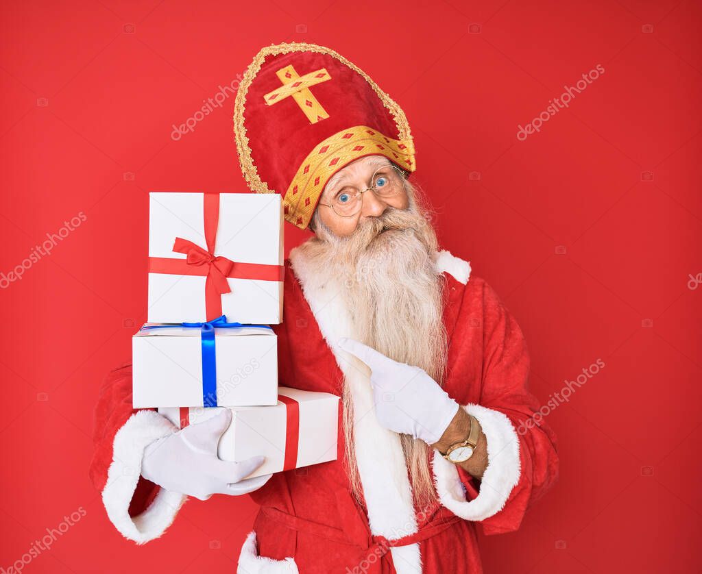 Old senior man with grey hair and long beard wearing saint nicholas costume holding presents smiling happy pointing with hand and finger 