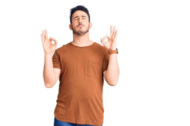 Günlük kıyafetler giyen genç İspanyol adam rahatla ve gözleri kapalı gülümse parmaklarıyla meditasyon hareketi yapıyor. yoga konsepti. 