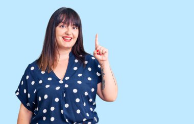 Genç ve cüsseli kadın gündelik kıyafetler giyer ve bir numaralı parmağını göstererek mutlu ve kendinden emin bir şekilde gülümser.. 