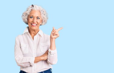 Yaşlı, gri saçlı, günlük kıyafetler giyen, yüzünde kocaman bir gülümseme olan, eli ve parmağı kameraya bakan bir kadın.. 