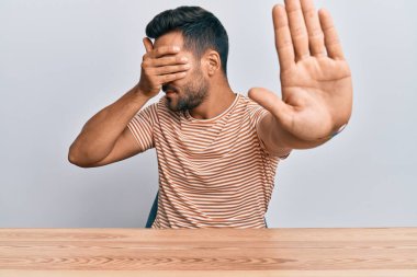 Yakışıklı İspanyol bir adam, gündelik giysiler giyip masaya oturur, gözlerini ellerle kapatır ve üzgün ve korku ifadeleriyle jestleri durdurur. utangaç ve olumsuz kavram. 