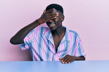 Gündelik giysiler giymiş genç Afro-Amerikan bir adam masaya oturmuş gülümsüyor ve elleri yüzüne gülüyor şaşkınlıktan gözleri kapatıyordu. kör kavram. 