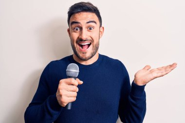 Beyaz arka plan üzerinde mikrofon kullanarak şarkı söyleyen sakallı yakışıklı genç şarkıcı başarıyı mutlu bir gülümsemeyle ve el kaldırarak kazanan ifadeyle kutluyor.