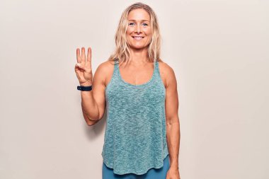Orta yaşlı beyaz kadın spor giysisi giyer ve üç numaralı parmaklarıyla gülümseyerek mutlu ve kendinden emin bir şekilde gösterir.. 