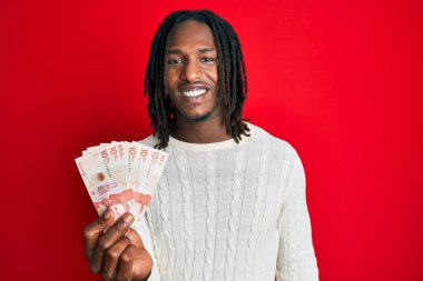 Saçları örgülü, elinde 10 Kolombiya pezosu olan Afro-Amerikan bir adam. Olumlu ve mutlu görünüyor. Dişlerini gösteren bir gülümsemeyle ayakta duruyor. 