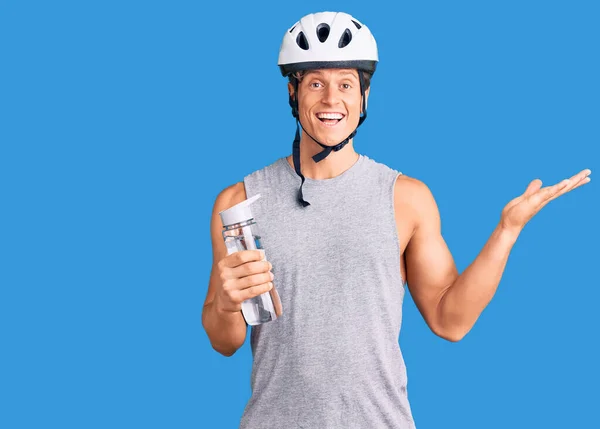 Young handsome man wearing bike helmet holding bottle of water celebrating victory with happy smile and winner expression with raised hands