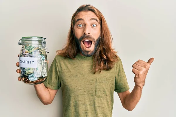 Attractive man with long hair and beard holding charity jar with money pointing thumb up to the side smiling happy with open mouth