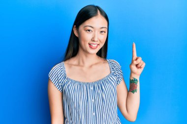 Gündelik çizgili tişört giyen Çinli genç bir kadın bir numaralı parmağını gösterip onu gösterirken kendinden emin ve mutlu bir şekilde gülümsüyor.. 