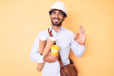 Kıvırcık saçlı yakışıklı genç adam ve ayı mimar şapkası takıyor inşaat projesi yapıyor ve kahve içiyor parmaklarıyla imza atıyor, arkadaşça gülümseyerek mükemmel bir sembol oluşturuyor. 