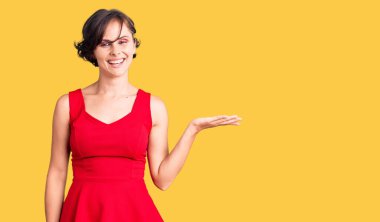 Kısa saçlı, rahat giyimli, kolsuz tişörtlü, neşeli bir şekilde gülümseyen ve avuç içi kameraya bakan güzel bir genç kadın.. 