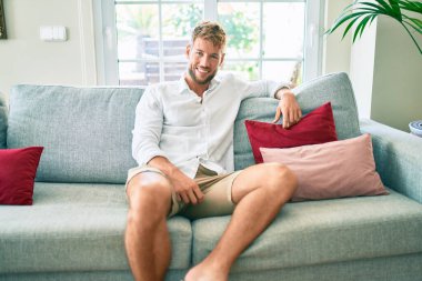 Yakışıklı beyaz adam mutlu bir şekilde gülümsüyor. Evdeki kanepede oturuyor.