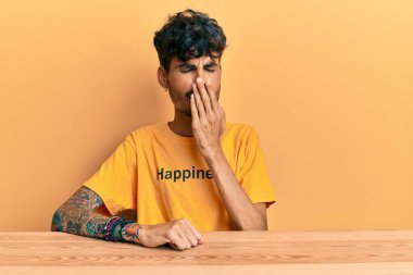 Üzerinde mutluluk mesajı olan tişört giyen genç İspanyol adam masada oturup ağzını eliyle kapatmaktan sıkılmış. Huzursuz ve uykulu.. 