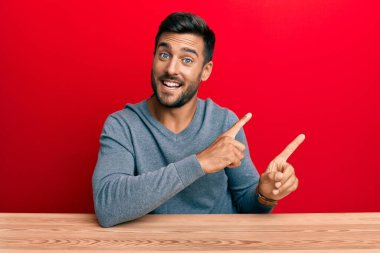 Yakışıklı İspanyol bir adam sıradan giyinmiş masaya oturmuş gülümsüyor ve kameraya bakıyor iki eliyle ve parmaklarıyla kenarı işaret ediyor.. 