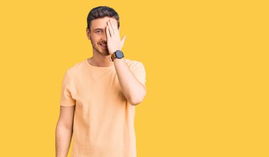 Ayılı yakışıklı genç adam gündelik sarı tişört giyiyor bir gözünü eliyle kapatıyor, yüzünde kendine güvenen bir gülümseme ve sürpriz bir duygu var.. 