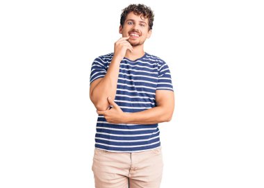 Kıvırcık saçlı, sıradan kıyafetler giyen, kameraya kendinden emin, kollarını kavuşturmuş ve çenesini kaldırmış gülümseyen genç yakışıklı bir adam. Olumlu düşünmek. 