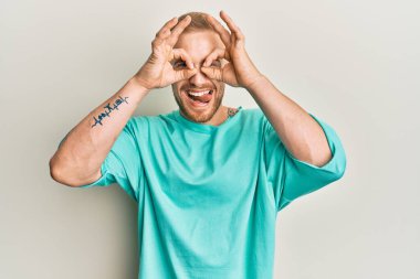 Kafkasyalı genç bir adam günlük kıyafetler giyiyor dürbün gibi jest yapıyor, dili dışarı çıkarıyor, gözleri parmaklarından bakıyor. çılgın ifade. 