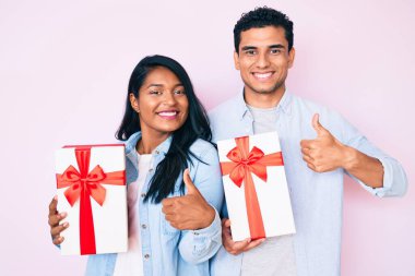 Güzel, genç bir çift yıldönümü kutlaması için hediye taşıyor mutlu ve pozitif gülümsüyor, başparmağıyla mükemmel ve onay işareti yapıyor. 