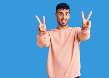 Gündelik giysiler giymiş, ağzı açık gülümseyen genç İspanyol adam zafer işareti yaparken parmaklarını gösteriyor. İki numara.. 