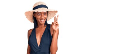 Mayo giyen, yaz şapkalı, gülümseyen, kameraya göz kırpan ve parmaklarıyla zafer işareti yapan Afro-Amerikan genç bir kadın. İki numara.. 