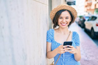 Genç ve güzel bir kız mutlu bir şekilde gülümsüyor. Akıllı telefon kullanıyor ve şehrin duvarına yaslanıyor.