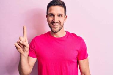 İzole edilmiş pembe arka planda dikilmiş tişört giyen genç yakışıklı bir adam bir fikir veya soruyla gülümsüyor ve mutlu bir yüzle işaret ediyor.