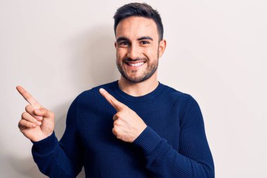 Sakallı, sıradan bir süveter giyen, beyaz arka planda duran, gülümseyen ve kameraya bakan iki eli ve parmakları yandan gösteren genç yakışıklı bir adam..