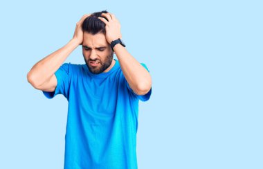 Sakallı, gündelik tişörtlü, baş ağrısı çeken, çaresiz ve stresli genç bir adam. Ağrı ve migren yüzünden. Eller başının üstünde.. 