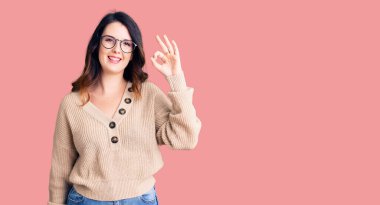 Güzel, genç, esmer bir kadın, günlük kıyafetler ve gözlük takıyor, gülümsüyor ve el ve parmaklarıyla 