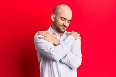 Zarif bir gömlek giyen genç yakışıklı kel adam kendine sarılıyor mutlu ve pozitif, kendinden emin gülümsüyor. Sevgi ve özgüven. 