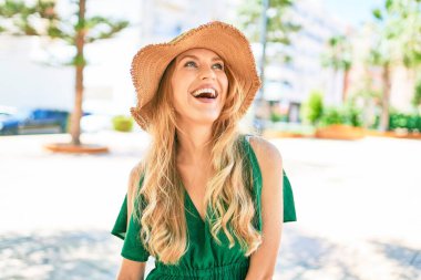 Yaz şapkası takmış, mutlu bir şekilde gülümseyen güzel sarışın bir kadın. Şehrin caddesinde yüzünde gülümsemeyle dikilirken.