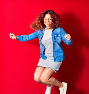 Genç, güzel, Latin bir kadın günlük kıyafetler giyip mutlu gülümsüyor. Yüzünde tebessümle zıplıyor, kırmızı arka planda yumruklarıyla kazanan jestler yapıyor.