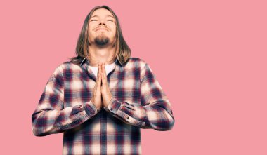 Uzun saçlı yakışıklı beyaz adam hippi tişörtü giyiyor, elleri bağlı yalvarıyor ve dua ediyor. Yüzünde umut ifadesi var. Çok duygusal ve endişeli. Yalvarıyor. 