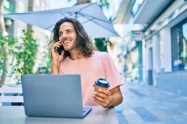 Genç İspanyol gülümseyen adam dizüstü bilgisayarla çalışırken ve kahve dükkanının terasında konuşurken mutlu oluyor..