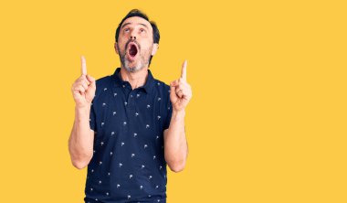 Orta yaşlı, polo giyen yakışıklı adam şaşırmış ve şaşırmış bir şekilde yukarı bakıyor ve parmaklarıyla işaret ediyor ve kollarını kaldırıyor.. 