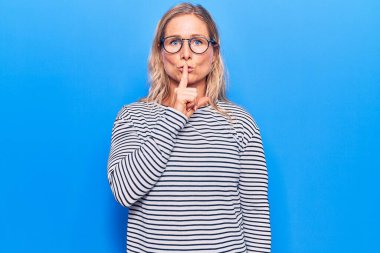 Orta yaşlı, beyaz, çizgili süveter ve gözlüklü, dudakları parmaklı, sessiz olmak isteyen sarışın kadın. sessizlik ve gizli konsept. 