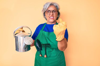 Bahçıvan önlüğü ve eldiven giyen yaşlı, beyaz saçlı bir kadın. Elinde sulama eldiveni olan. Öfkeyle bağıran, öfkeyle bağıran ve el kaldıran. 