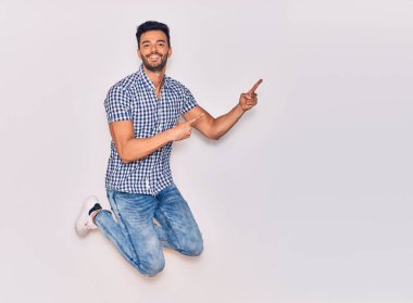 Genç, yakışıklı, sıradan kıyafetler giyen, mutlu gülümseyen bir İspanyol. Yüzünde tebessümle sıçrayıp, izole beyaz arka planda parmaklarını yana doğru gösteriyor.