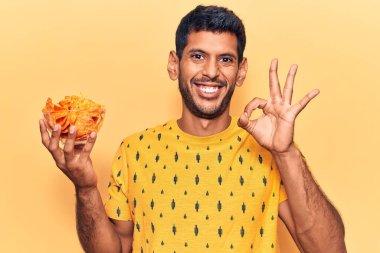 Elinde patates cipsi tutan genç latin adam parmaklarıyla imza atıyor, gülümseyerek mükemmel bir sembol oluşturuyor. 