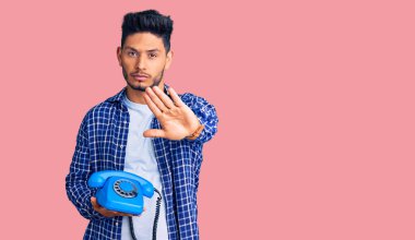 Yakışıklı Latin Amerikalı genç adam elinde klasik bir telefonla dur işareti yapıyor ciddi ve kendinden emin bir ifadeyle, savunma jesti yapıyor. 