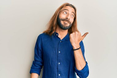 Uzun saçlı ve sakallı çekici bir adam gündelik giysiler giyip baş parmağını kaldırıyor ve ağzı açık gülümsüyor. 